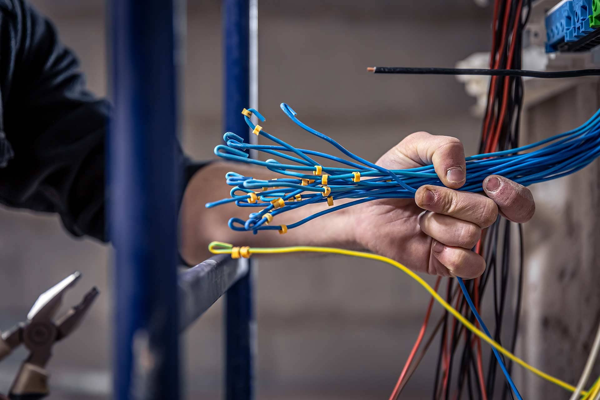 Iron Fibre Networks A person holds multiple blue electrical wires with yellow connectors, preparing for installation. Nearby are other wires and a pair of pliers, suggesting a workspace set up for Fibre Optic Cable Solutions & Services. Fibre Optic Cable Solutions & Services