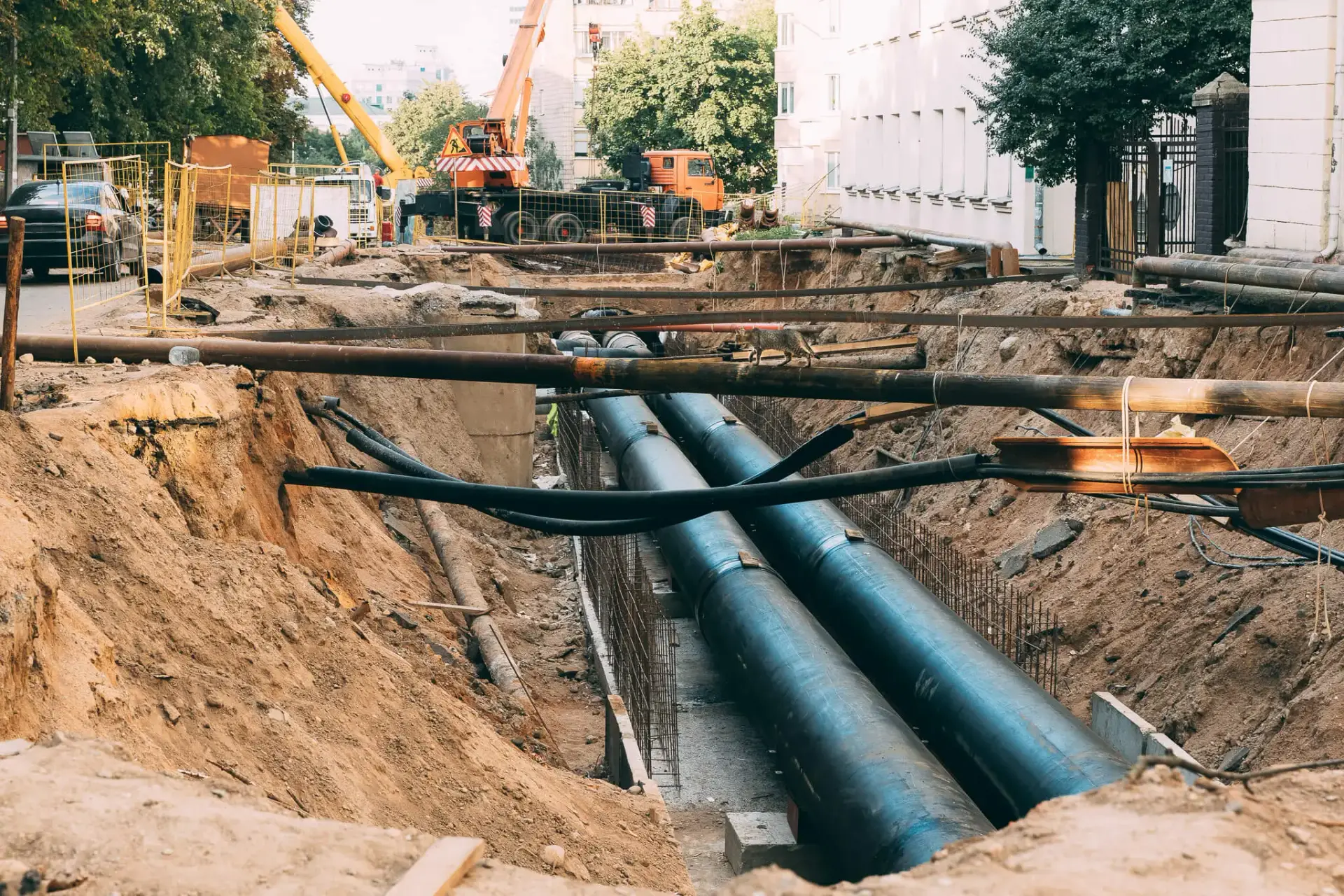 Iron Fibre Networks Large exposed pipes run through a deep trench at a construction site, where Fibre Optic Cable Solutions & Services are being implemented; cranes, fencing, and buildings are visible in the background. Fibre Optic Cable Solutions & Services