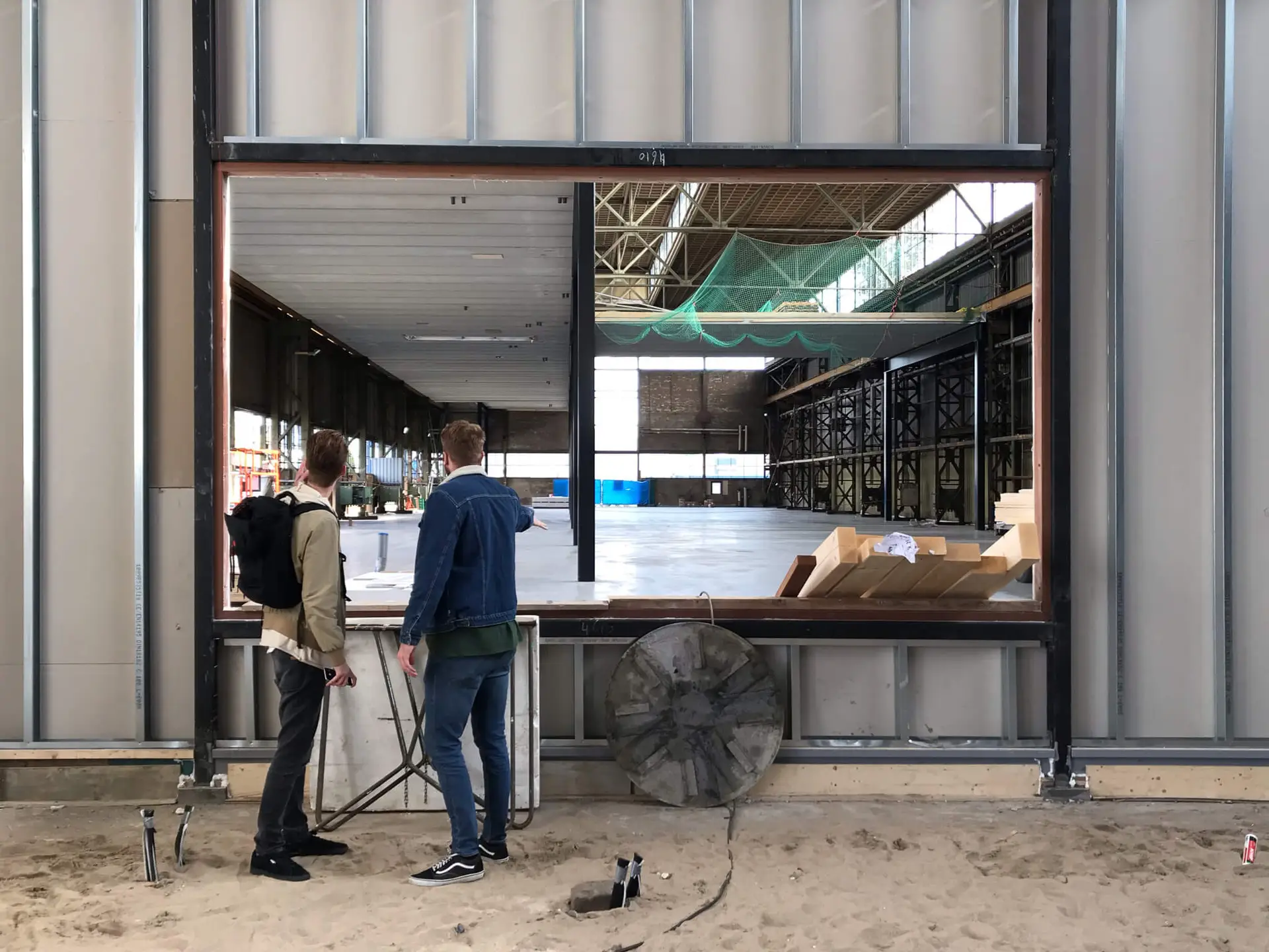 Iron Fibre Networks Two people stand on sandy ground, looking through a large open window frame into an unfinished industrial building with metal walls and construction materials inside, where Fibre Optic Cable Solutions & Services are set to enhance the infrastructure. Fibre Optic Cable Solutions & Services