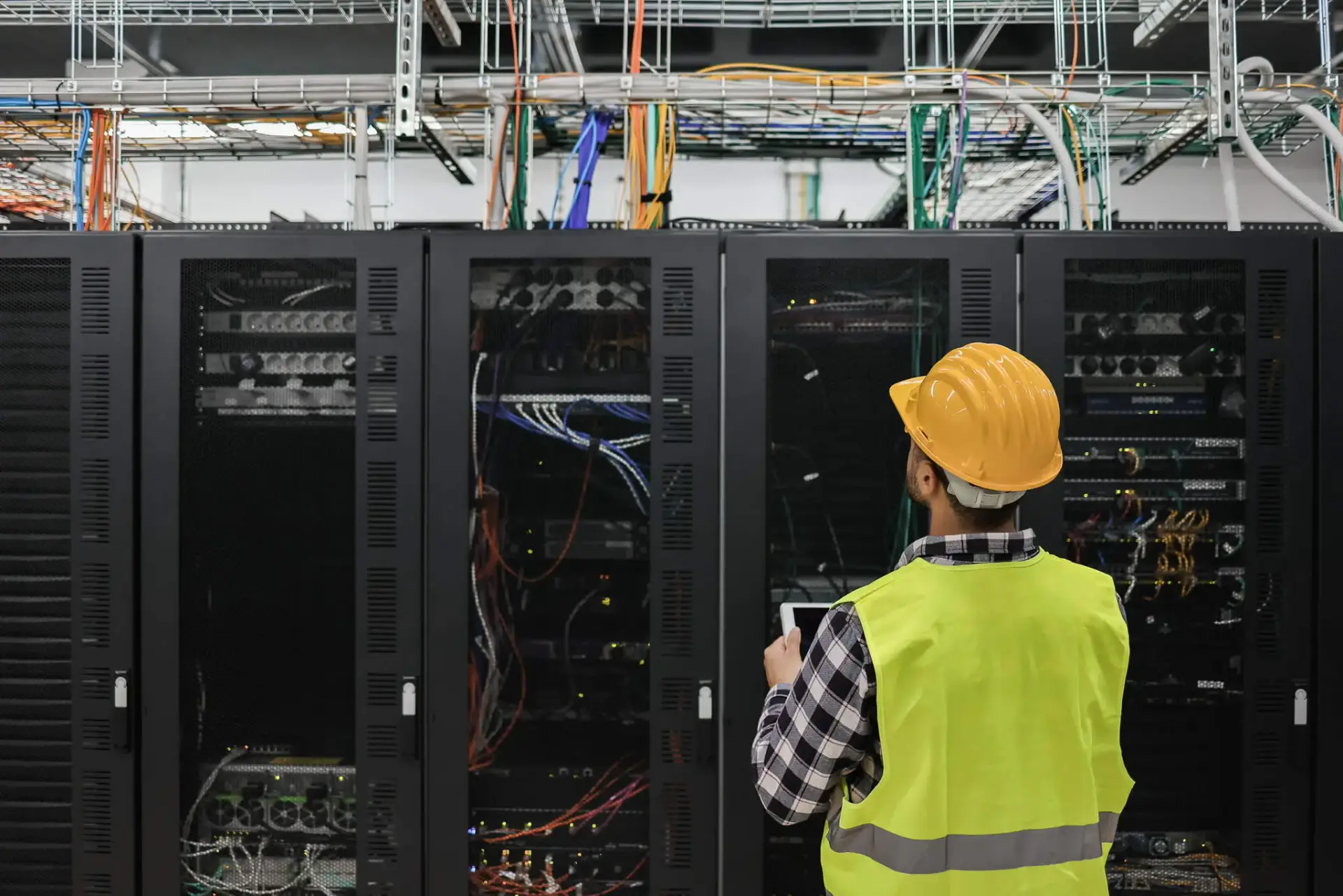 Iron Fibre Networks A worker in a yellow safety vest and hard hat inspects server racks with visible cables in a data center, ensuring the integrity of Fibre Optic Cable Solutions & Services. Fibre Optic Cable Solutions & Services